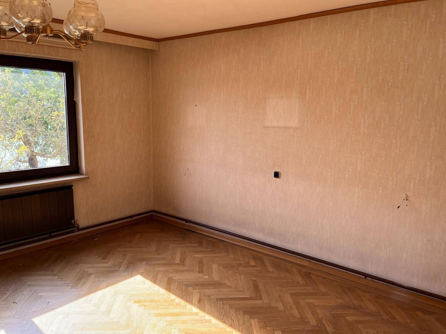 Leeres Zimmer mit Fischgrätparkett, Fenster und renovierungsbedürftigen Wänden.