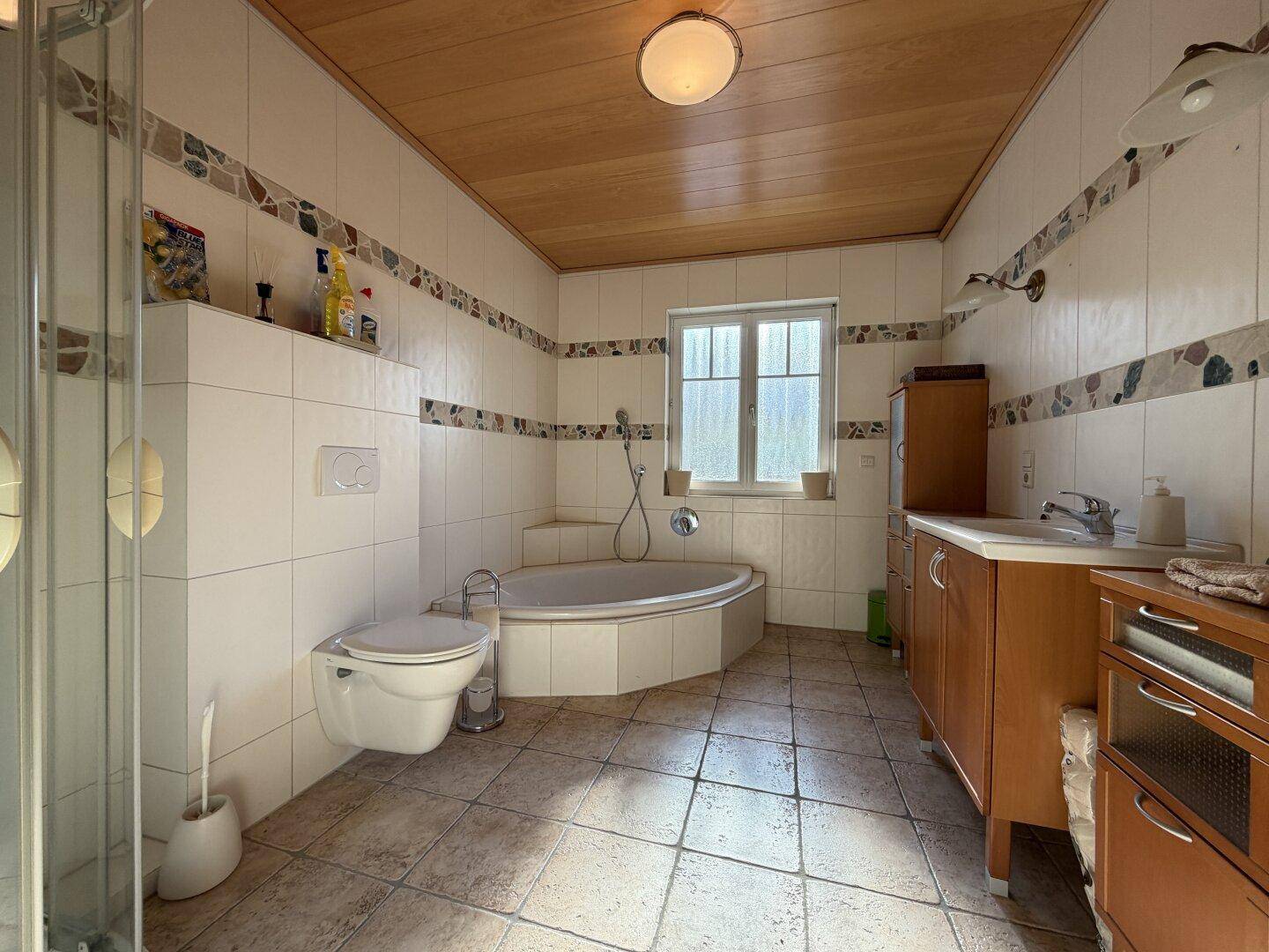 Badezimmer mit Eckbadewanne, Dusche, WC, Holzdecke und Fenster für Tageslicht.
