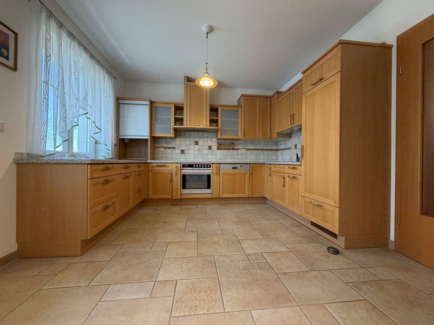 Geräumige Küche mit hellen Holzschränken, Fliesenboden und Fenster mit Vorhängen.
