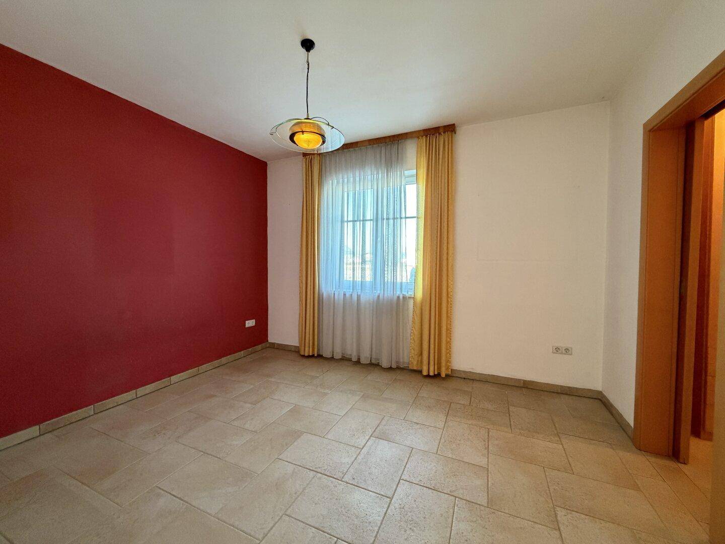 Leerer Raum mit roter Akzentwand, Fliesenboden und einem Fenster mit gelben Vorhängen.