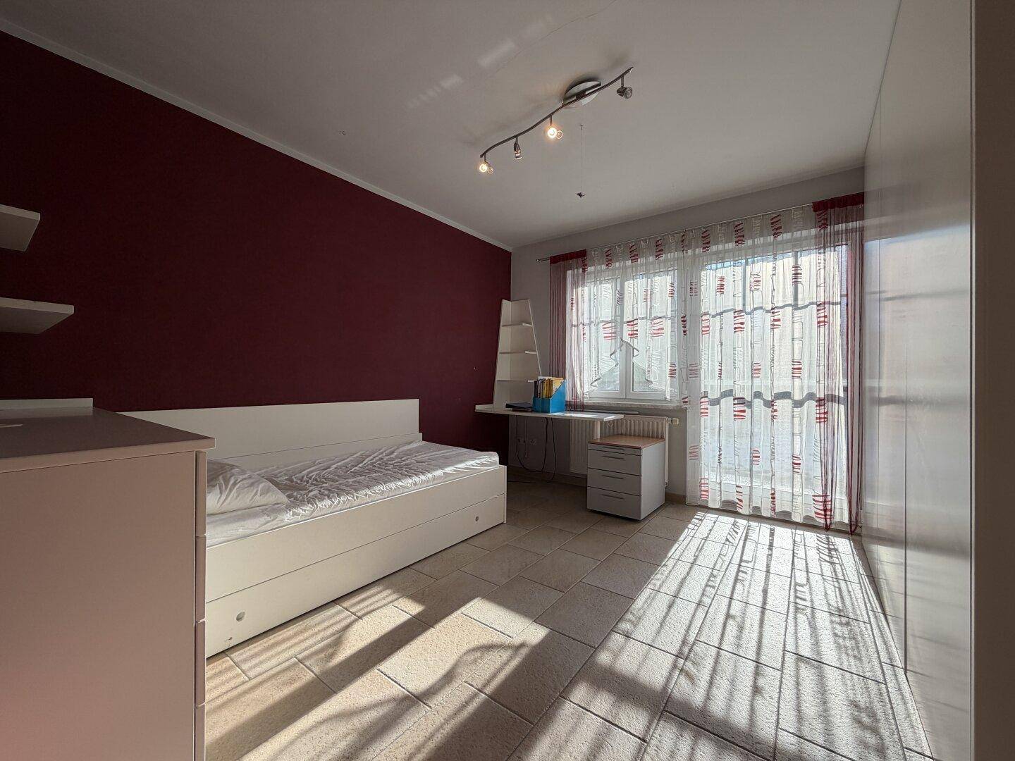 Helles Schlafzimmer mit Einzelbett, großem weißen Kleiderschrank und roter Akzentwand.
