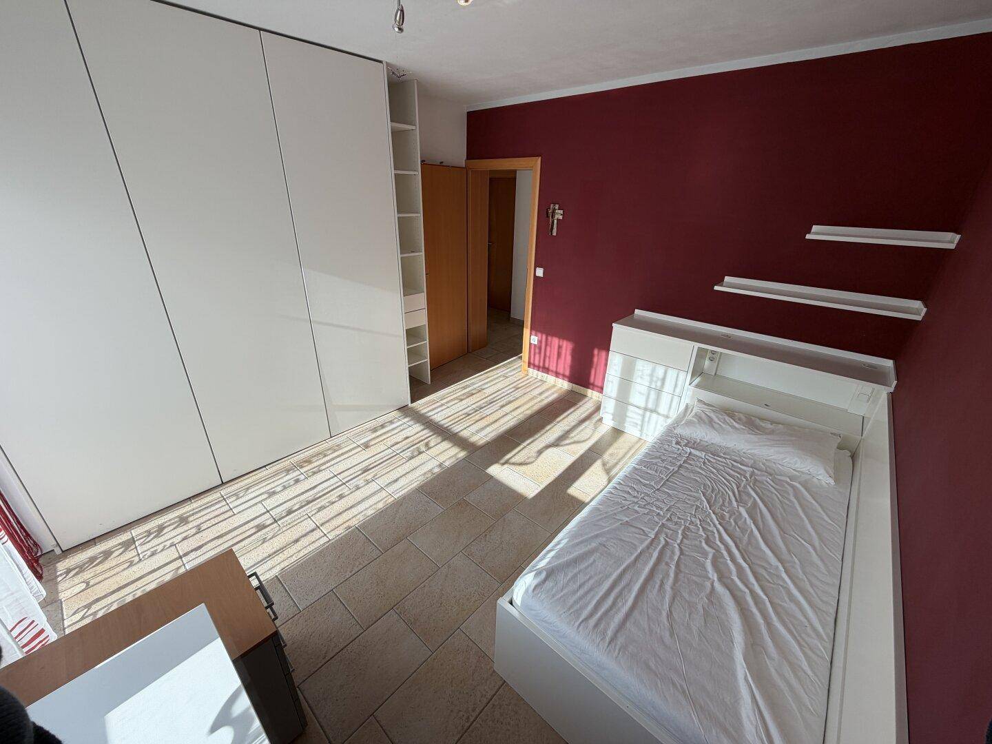 Schlafzimmer mit Einzelbett, weißen Schränken und roter Wand, durchflutet von natürlichem Licht.