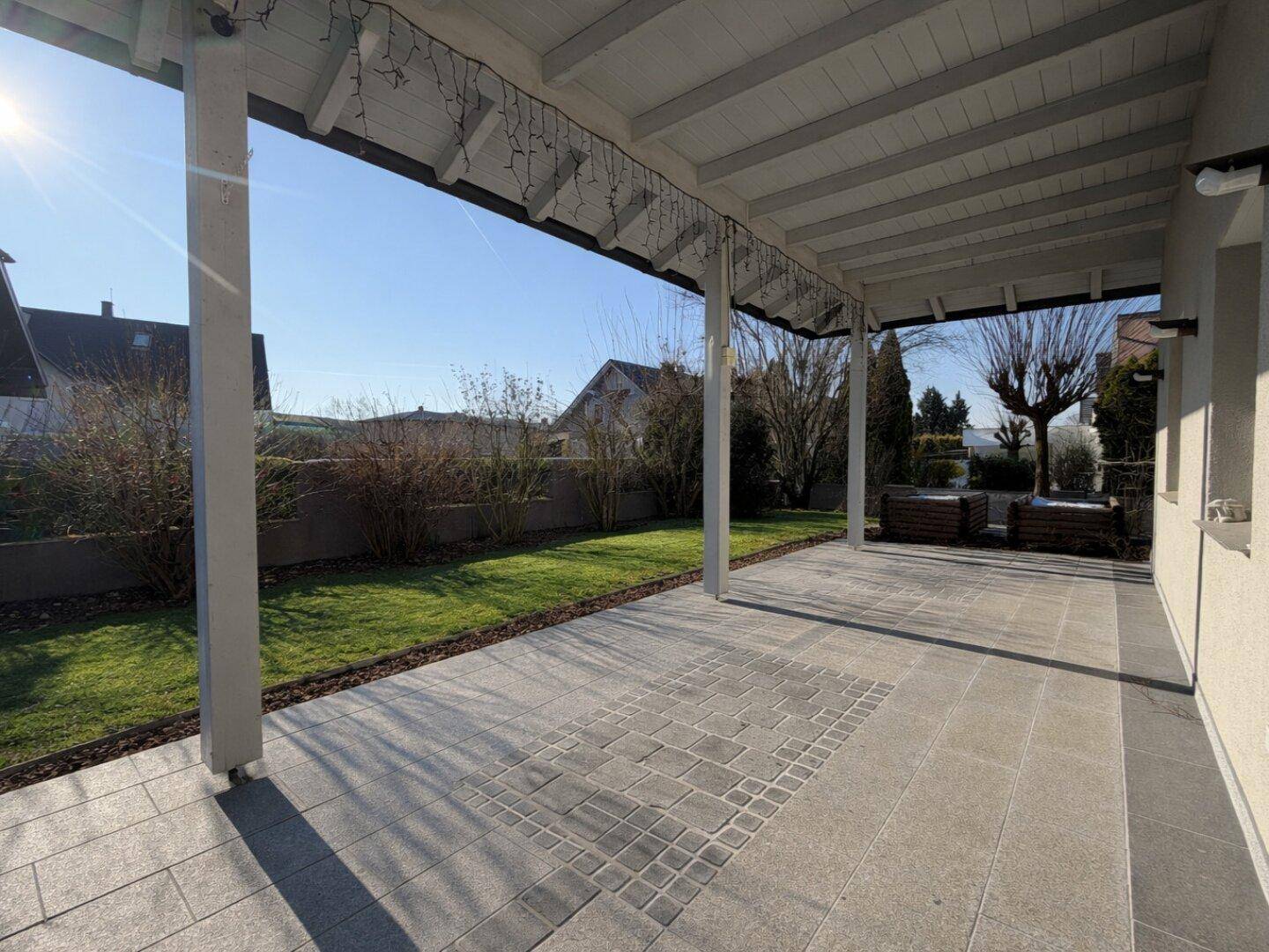 Geräumige überdachte Terrasse mit Pflastersteinen und Blick auf den grünen Garten.