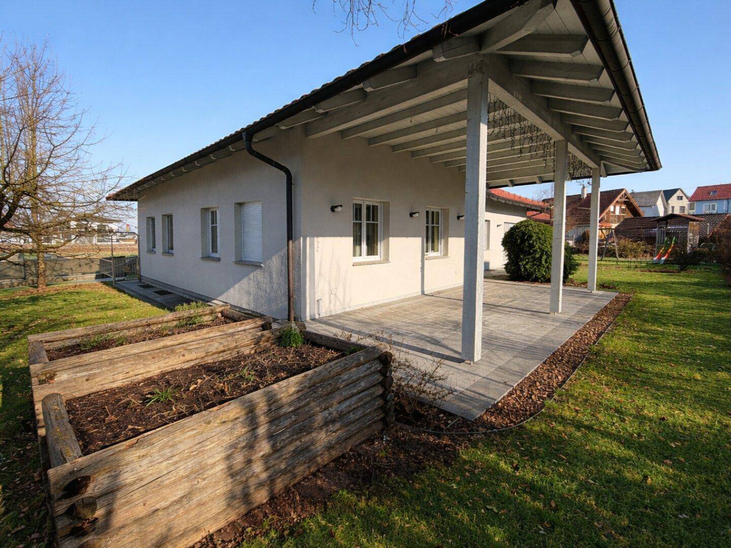 Überdachte Terrasse mit Holzbalken und erhöhten Hochbeeten im sonnigen Gartenbereich.