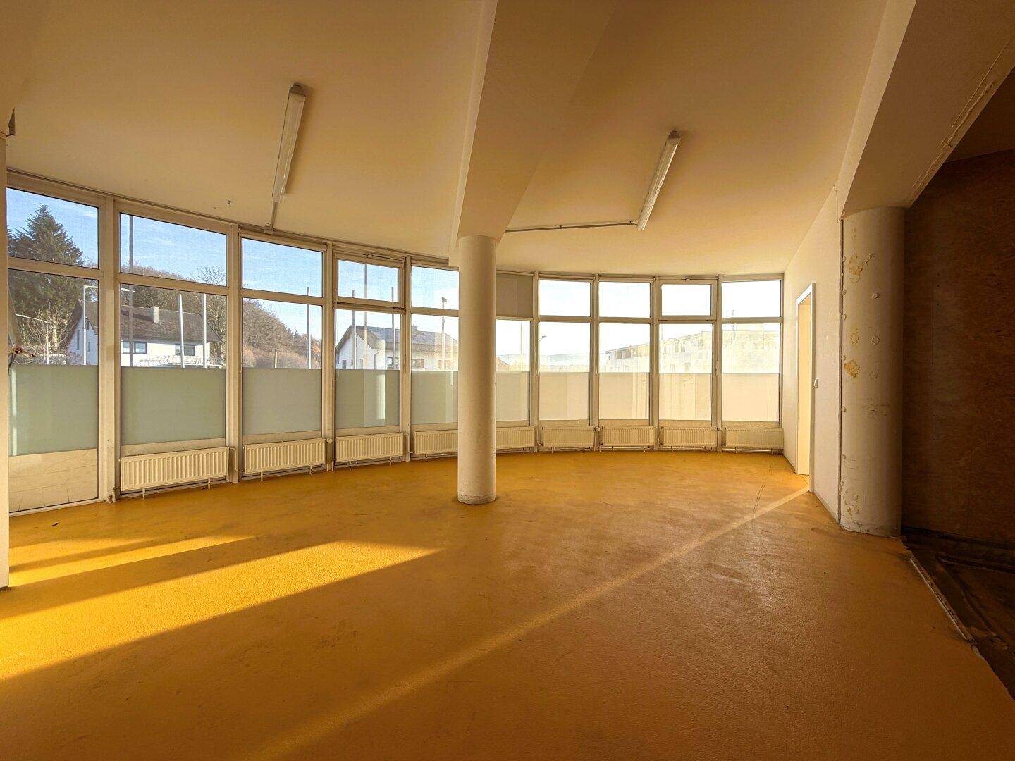 Großer, leerer Gewerberaum mit vielen Fenstern, die viel Tageslicht hereinlassen und einen Blick nach draußen bieten.