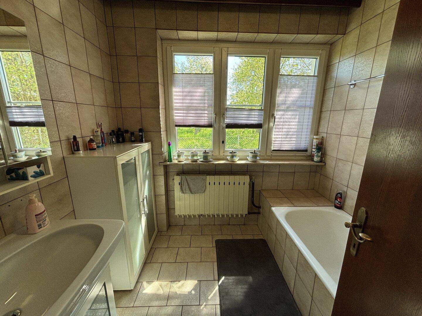 Badezimmer mit beigefarbenen Fliesen, Badewanne und Fenster für Tageslicht.