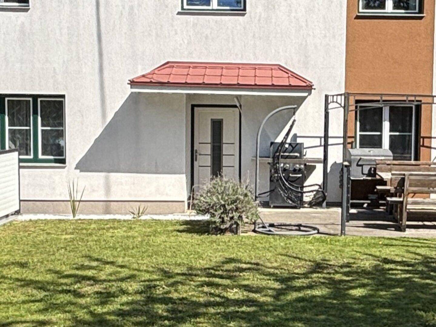 Eingangsbereich des Hauses mit rotem Vordach, weißer Tür und einem kleinen Rasenstück mit Grill.