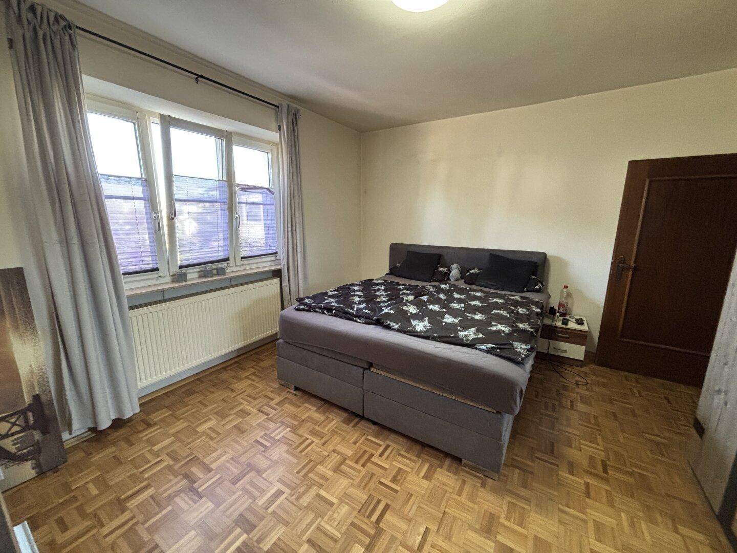 Schlafzimmer mit Doppelbett, Parkettboden und Fenster mit Jalousien.