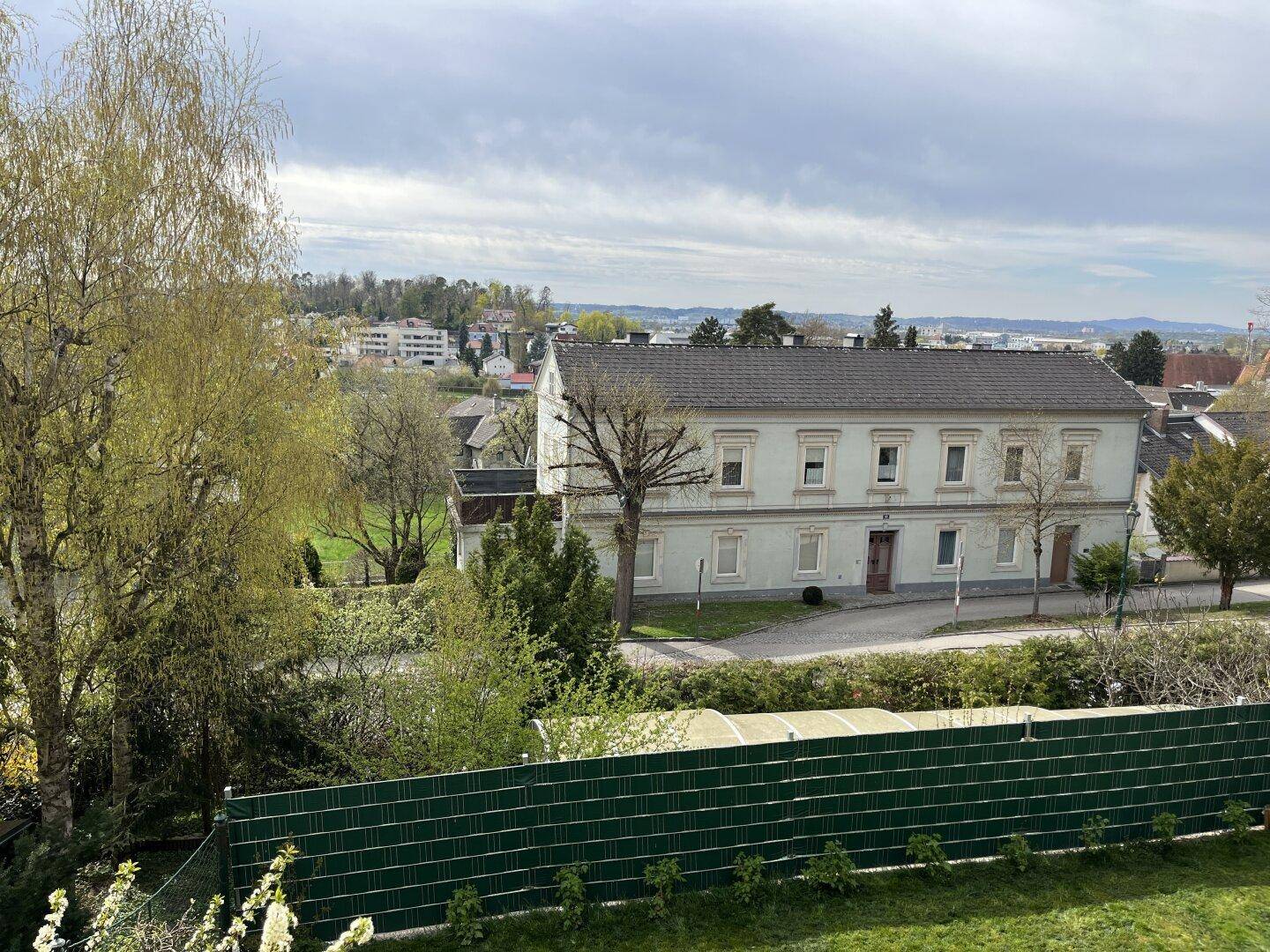 Weitläufiger Blick über eine grüne Wohngegend mit einem historischen Gebäude und hügeliger Landschaft im Hintergrund.