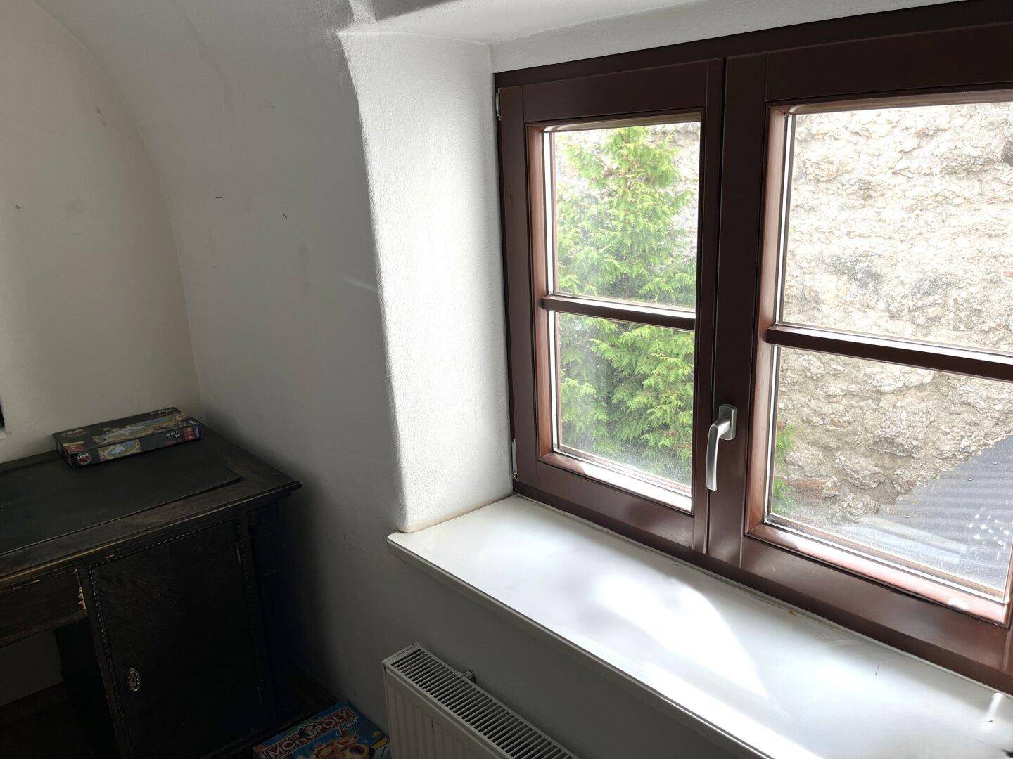 Fenster mit Blick auf eine Steinmauer und grüne Vegetation im Außenbereich.