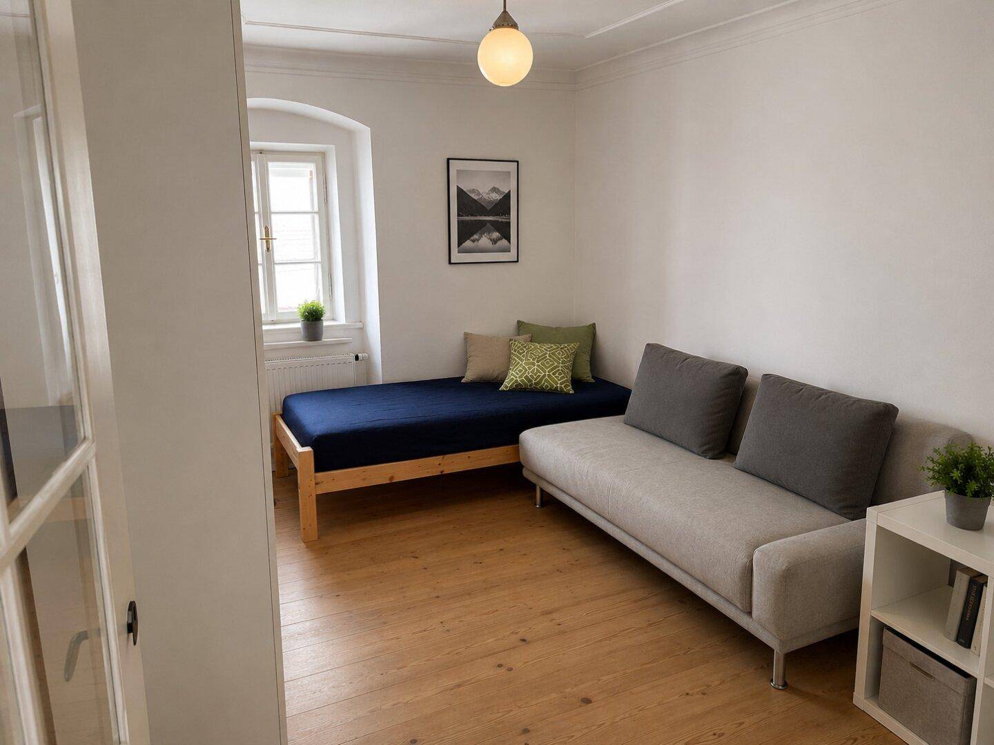 Helles Schlafzimmer mit Einzelbett, Sofa und Holzboden, ideal als Gästezimmer.