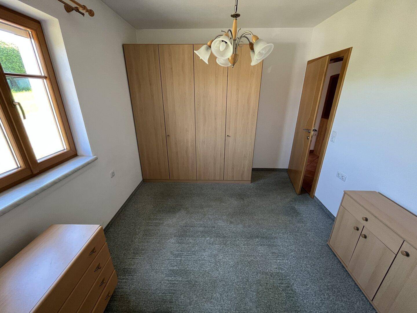 Schlafzimmer mit Teppichboden, großem Holzschrank und einem Fenster für Tageslicht.