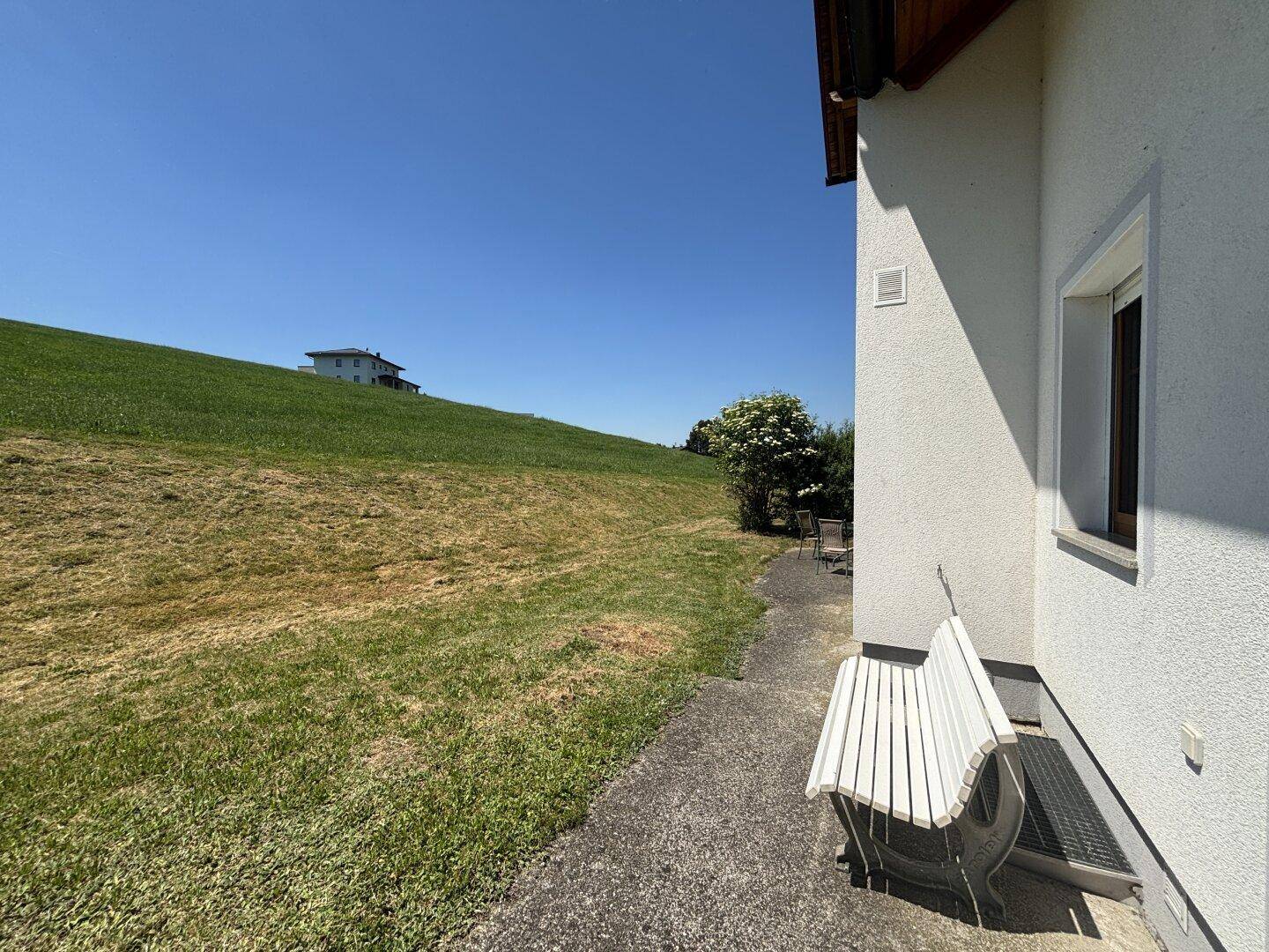 Außenbereich neben dem Haus mit einer weißen Bank und Blick auf eine grüne Hügellandschaft.