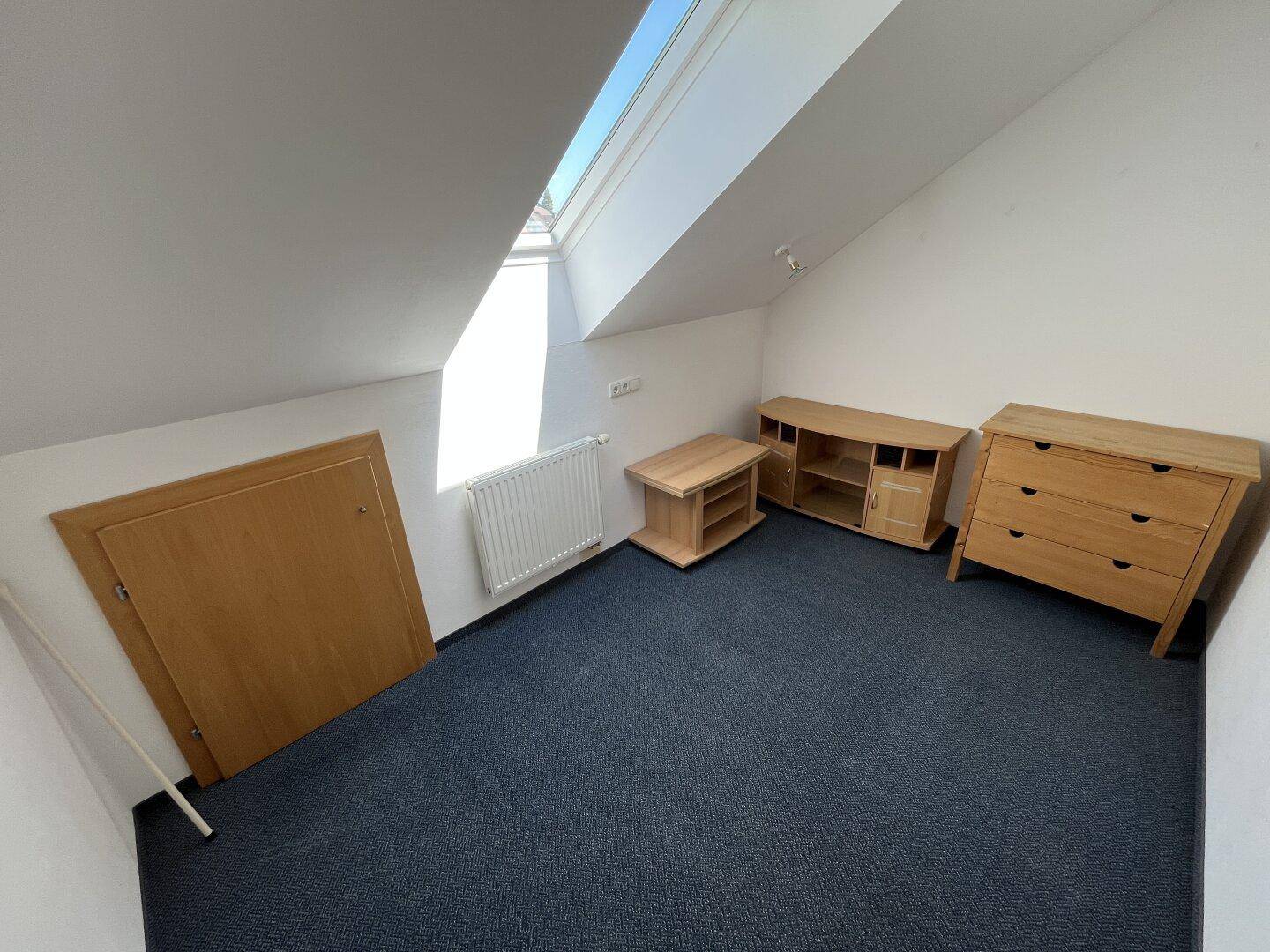 Dachschlafzimmer mit Teppichboden, Dachfenster und einfacher Möblierung.