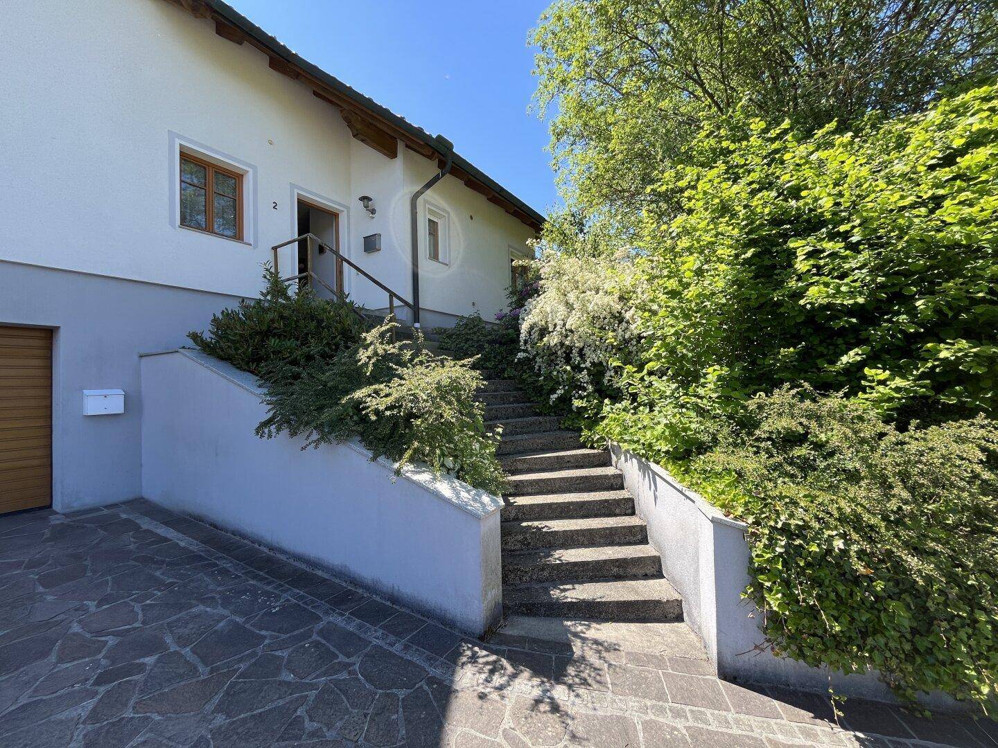 Eingangsbereich des Hauses mit Steintreppe, umgeben von üppigem Grün und Sträuchern.