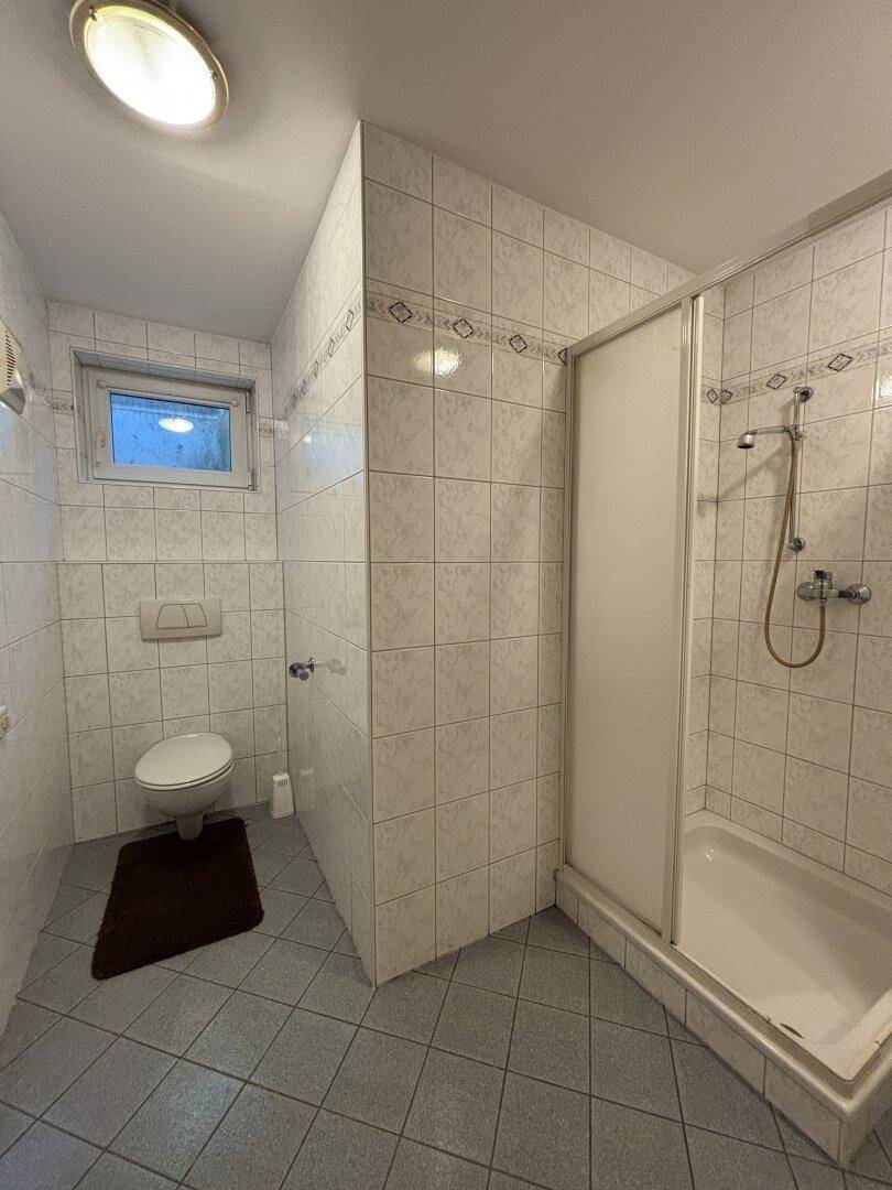 Gefliestes Badezimmer mit Toilette, Duschkabine und einem kleinen Fenster für natürliche Belüftung.