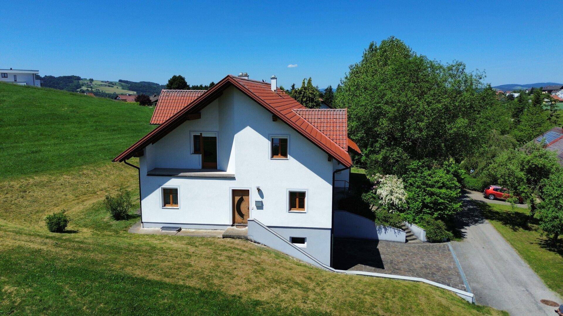 Freistehendes Haus auf einem grünen Hügel mit rotem Dach und umgeben von Bäumen unter blauem Himmel.