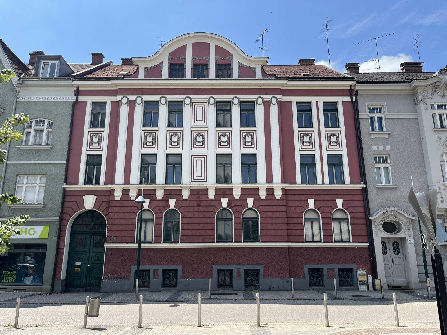 Historische Fassade eines Mehrfamilienhauses mit dekorativen Elementen und roten Akzenten an einer Straße.