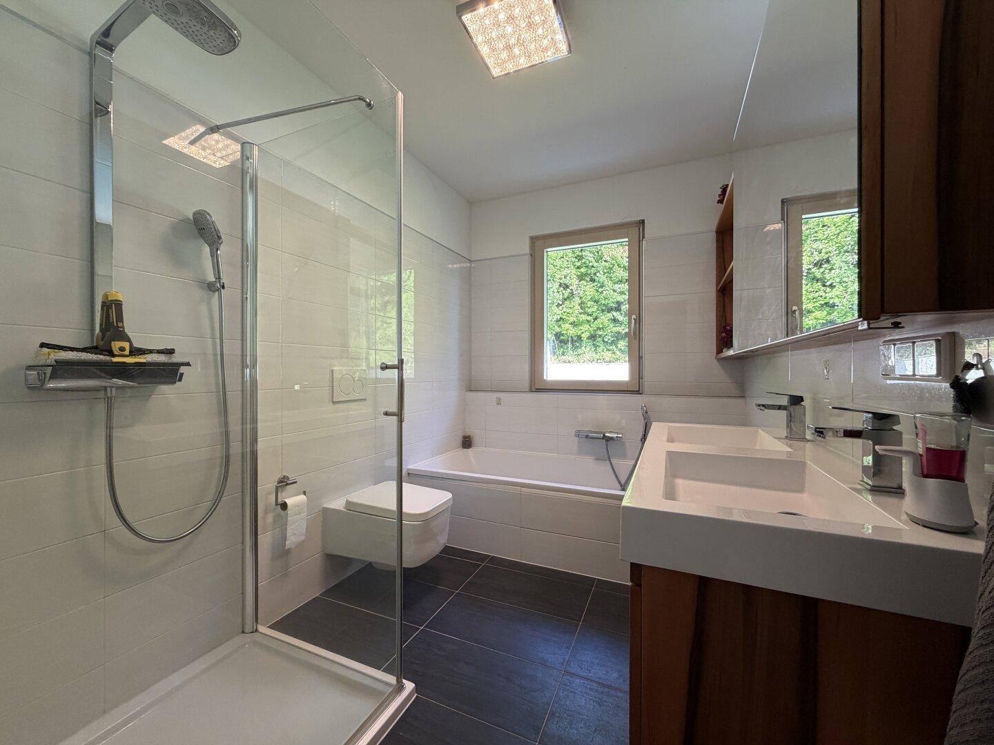 Modernes Badezimmer mit Doppelwaschbecken, Badewanne, Dusche und Fenster mit Blick ins Grüne.