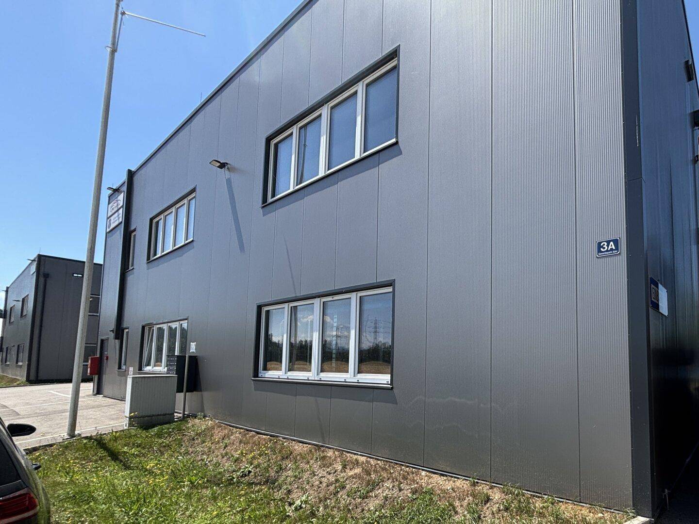 Seitenansicht eines modernen Industriegebäudes mit grauer Fassade und mehreren großen Fenstern.