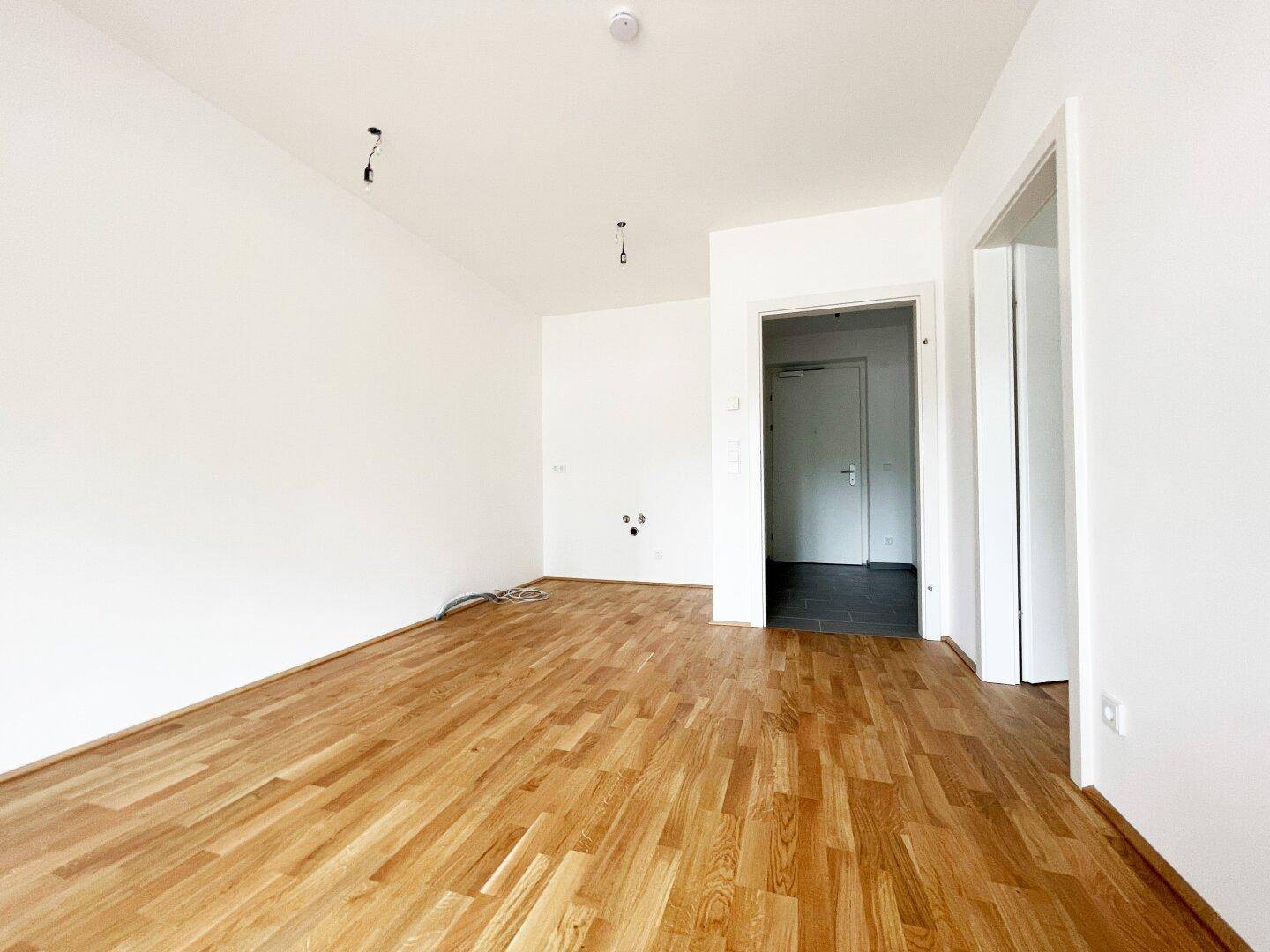 Geräumiges, leeres Wohnzimmer mit hellem Parkettboden und Blick auf den Eingangsbereich.