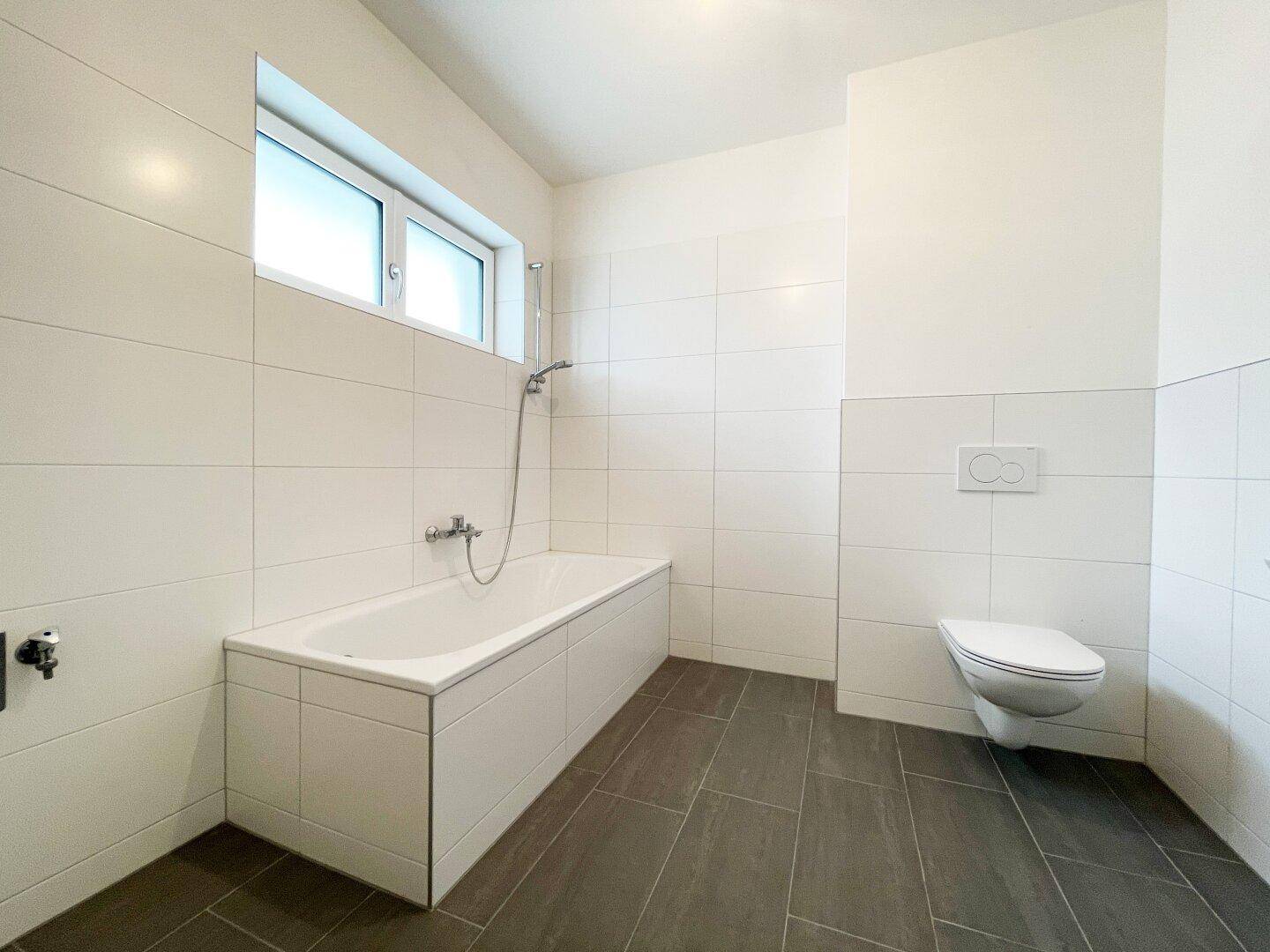 Modernes Badezimmer mit Badewanne, integrierter Duschfunktion, Wand-WC und großen weißen Fliesen.
