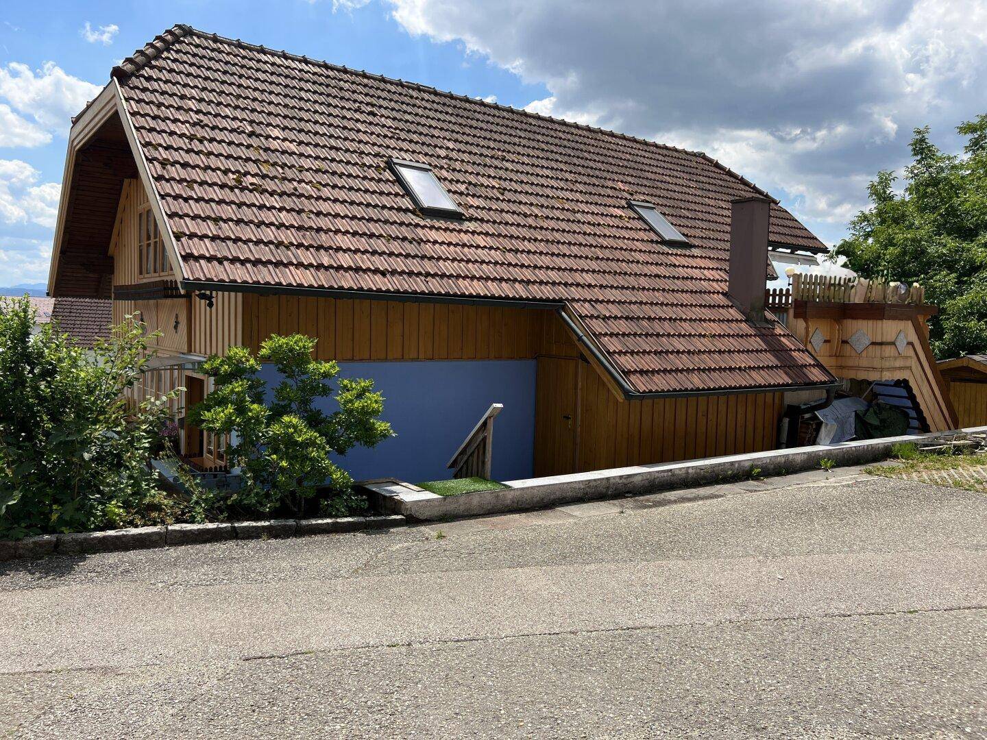 Seitenansicht eines Holzhauses mit Ziegeldach, Dachfenstern und blauer Sockelwand.