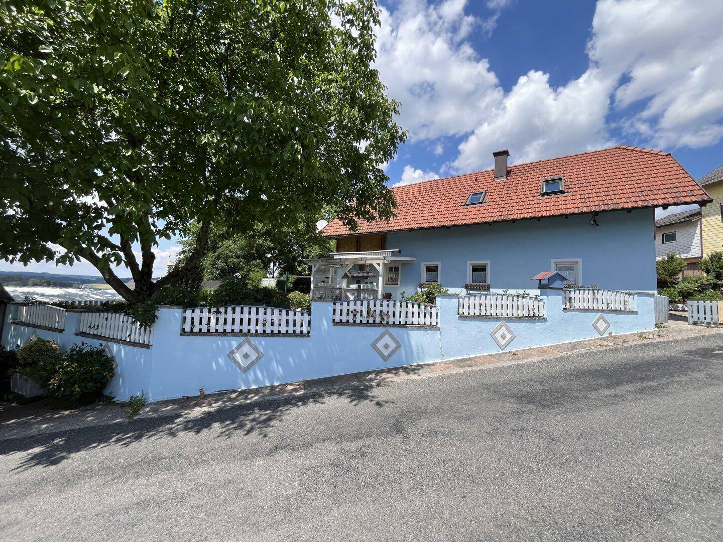Rückseite des Hauses mit blauer Fassade, weißem Zaun und Terrasse unter einem Baum.