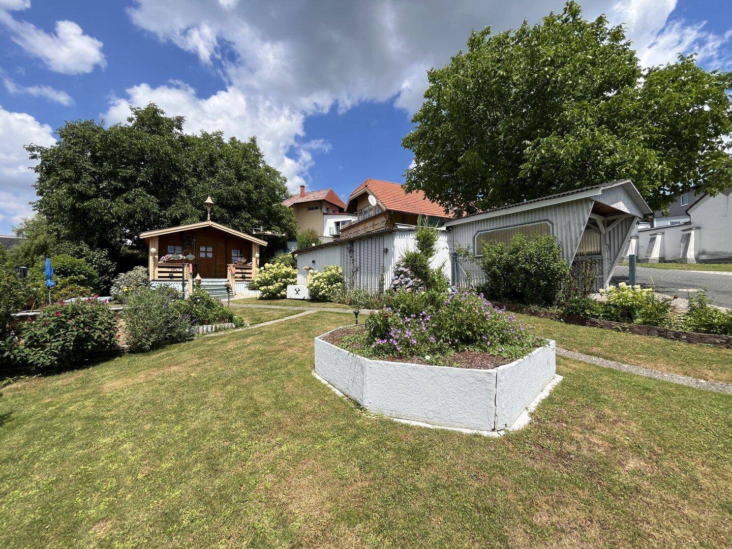 Weitläufiger Gartenbereich mit Rasenfläche, Blumenbeeten und Gartenhaus im Hintergrund.