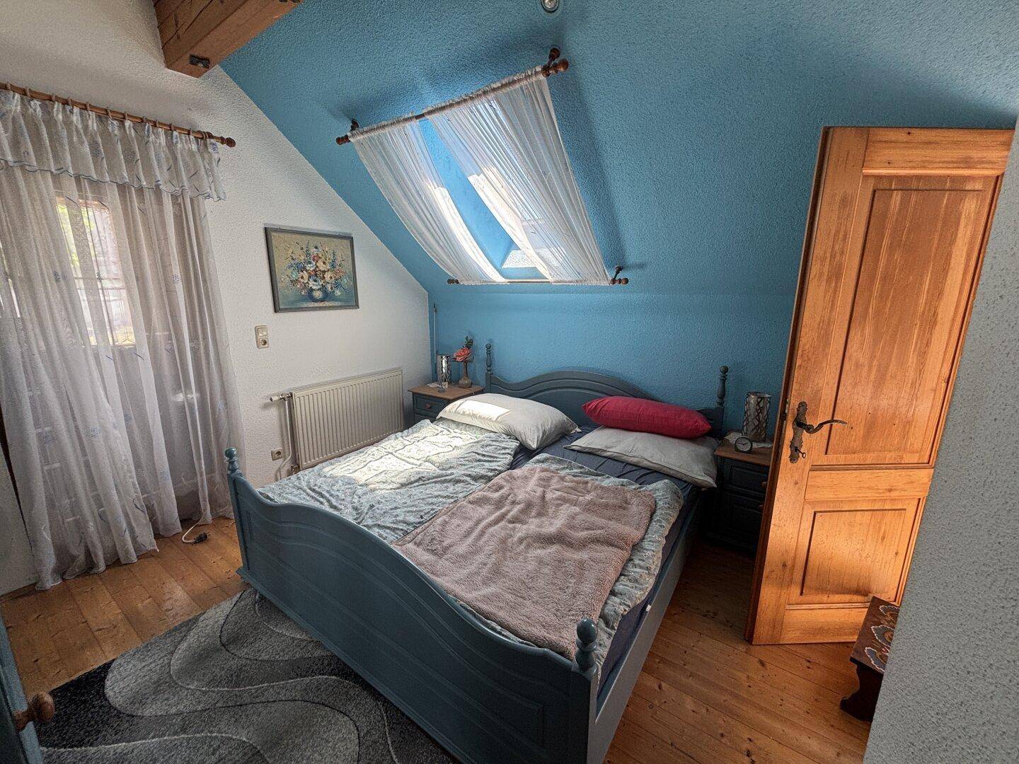 Schlafzimmer im Dachgeschoss mit Holzboden, blauer Wand und Dachfenster.