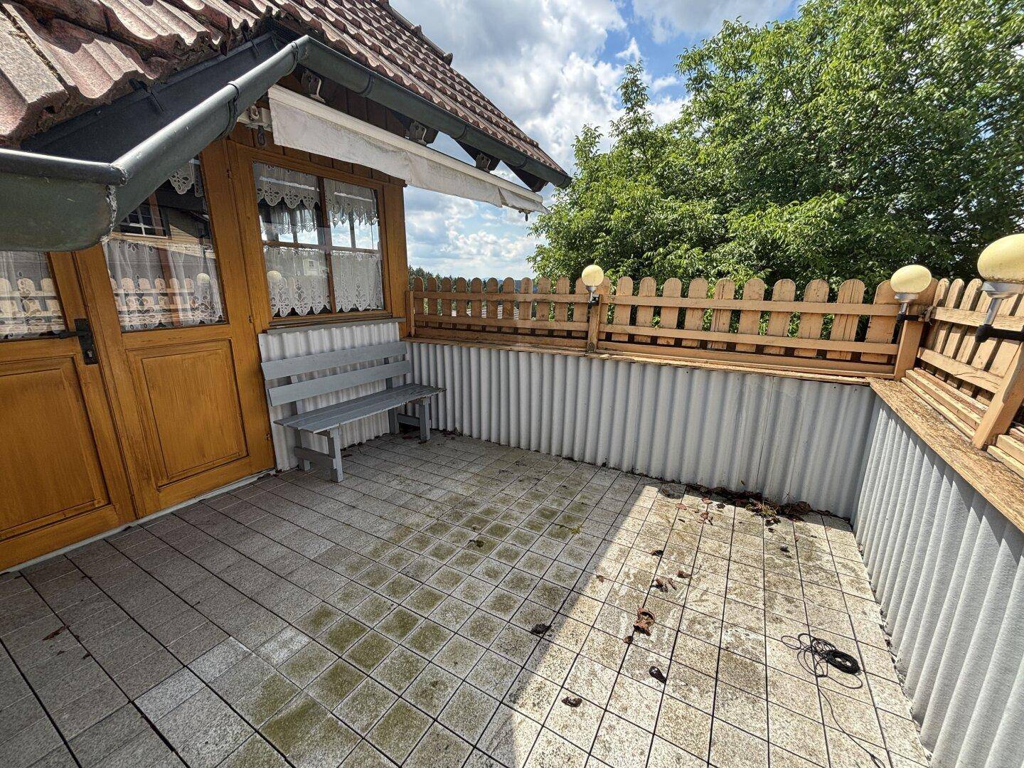 Geflieste Terrasse mit Holzbank und Holzzaun, umgeben von Bäumen.