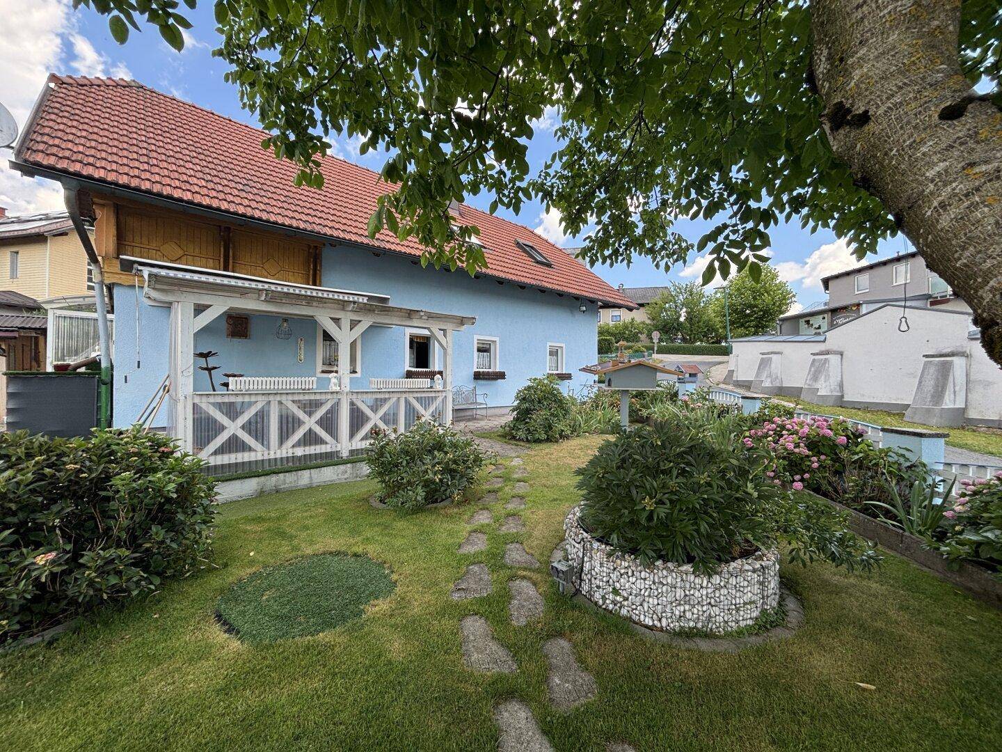 Gepflegter Garten mit Rasen, Steinpfad und blauen Hausfassade unter Bäumen.