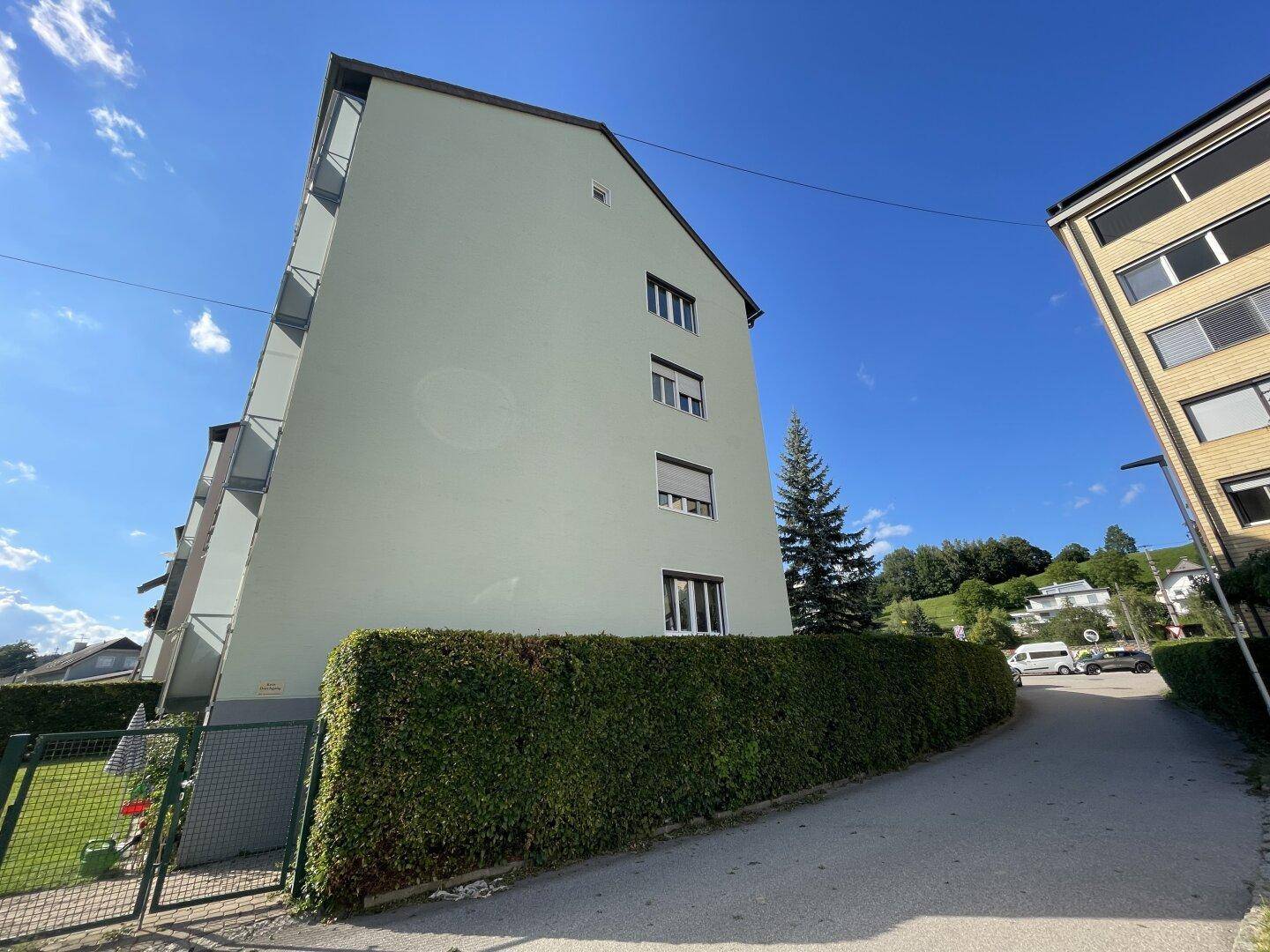 Seitenansicht eines Wohnhauses mit grüner Hecke und Straße im Hintergrund unter blauem Himmel.