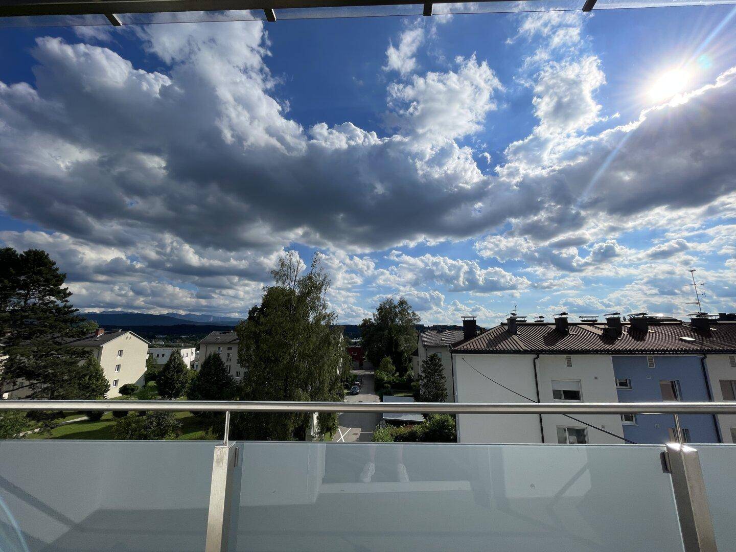 Heller Balkon mit Glasgeländer und weitem Blick über die Dächer und Berge bei Sonnenschein.