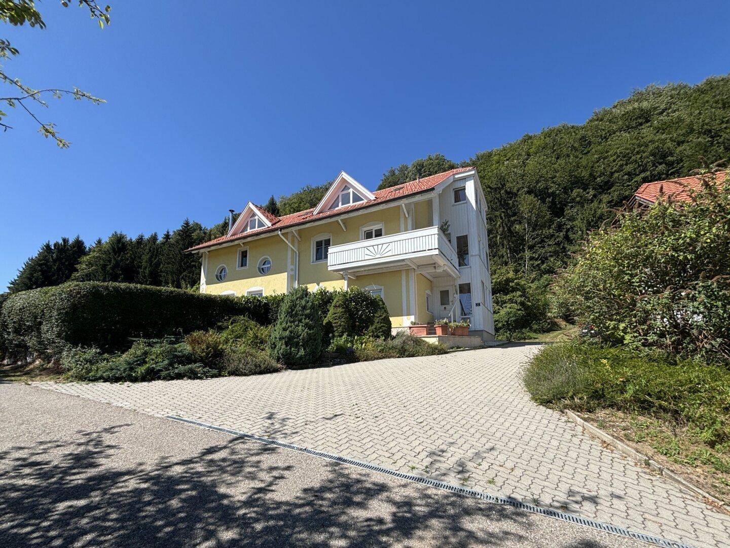 Gelbes Wohnhaus mit weißem Balkon, umgeben von Bäumen und Natur unter blauem Himmel.