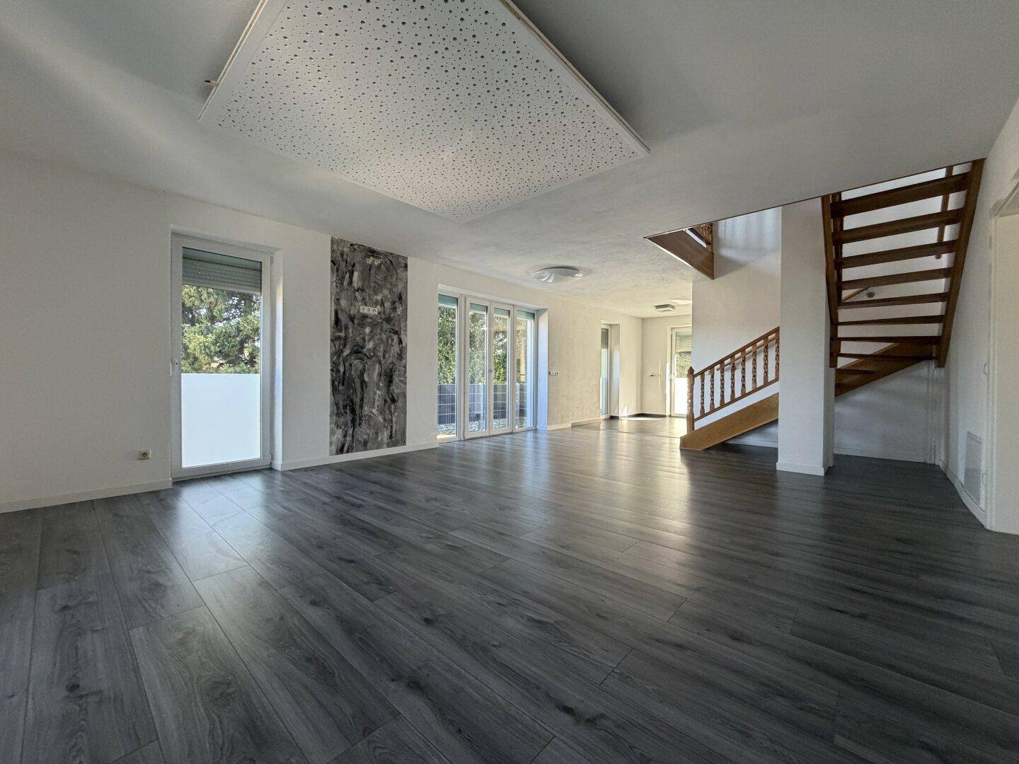 Heller Wohnbereich mit dunklem Laminatboden, großen Fenstern und Zugang zur Terrasse.