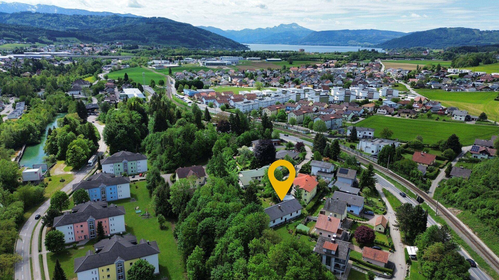 Panoramische Luftaufnahme einer Ortschaft mit See und Bergen im Hintergrund, die Lage markierend.