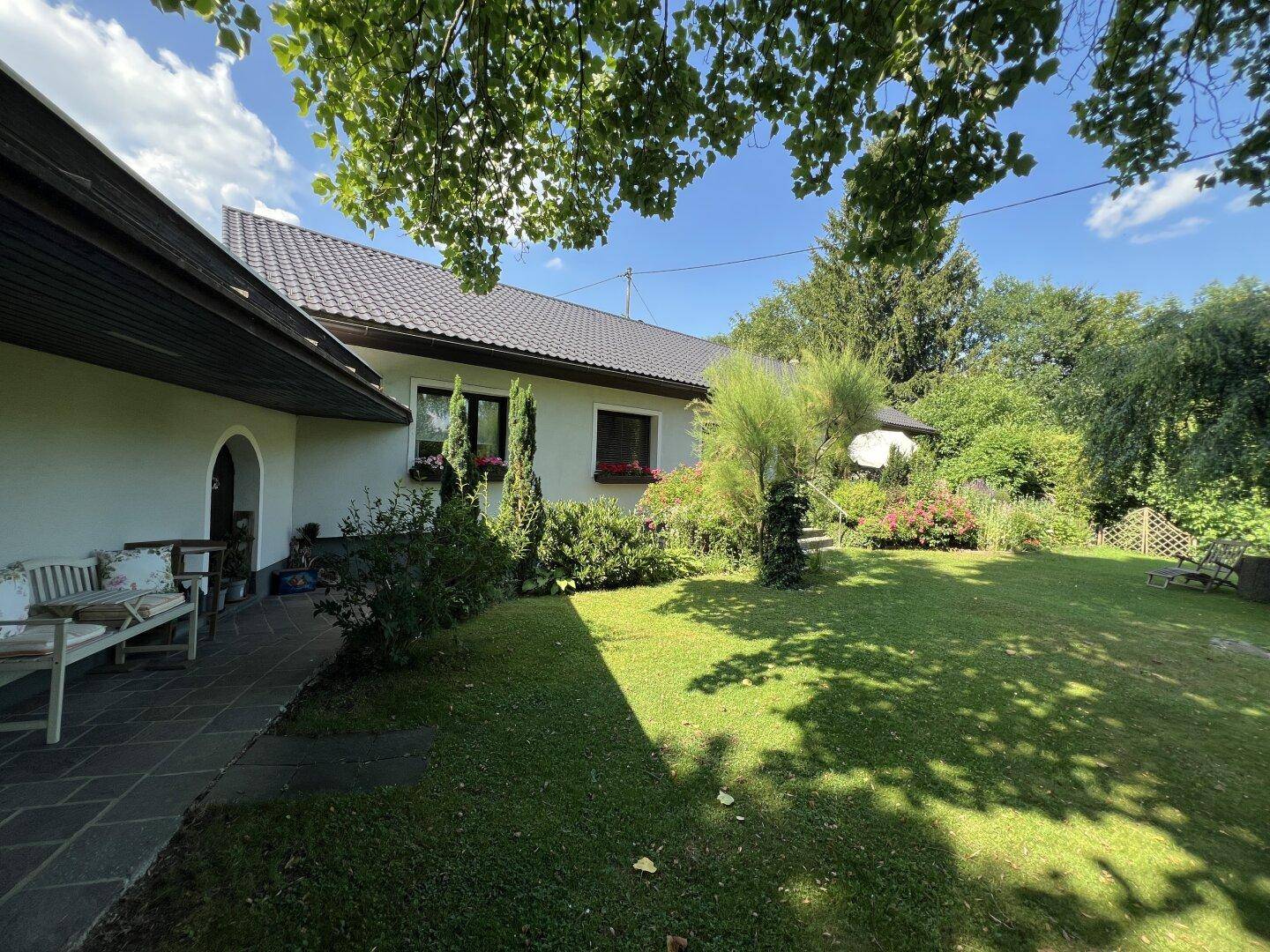 Seitlicher Blick auf das Haus mit gepflegtem Rasen, schattenspendenden Bäumen und einer kleinen Sitzgelegenheit.