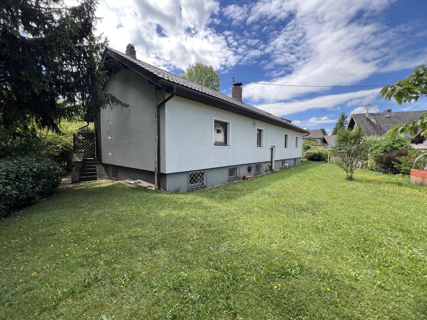 Rückseite des Hauses mit großem, sonnigen Rasenbereich und einfacher Fassade unter blauem Himmel.
