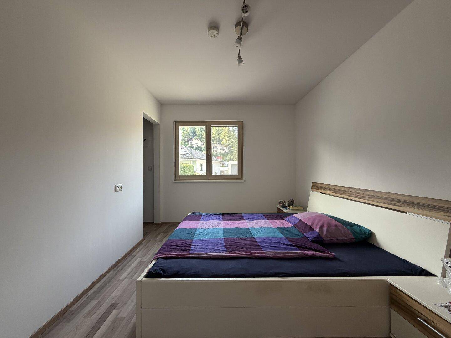 Schlafzimmer mit Doppelbett, Laminatboden und Fenster, das viel Tageslicht hereinlässt.