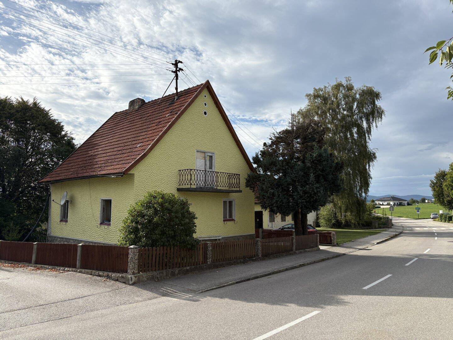 Gelbes Einfamilienhaus mit Satteldach, kleinem Balkon und ländlicher Umgebung unter bewölktem Himmel.