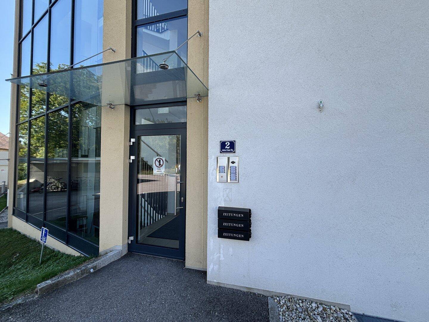 Moderner Eingangsbereich des Gebäudes mit Glasvordach und Gegensprechanlage.
