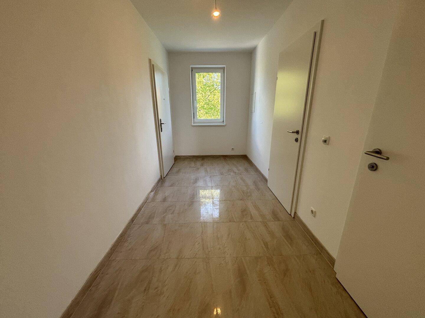 Leerer Flur mit hellen Fliesen und einem Fenster, das Tageslicht hereinlässt.