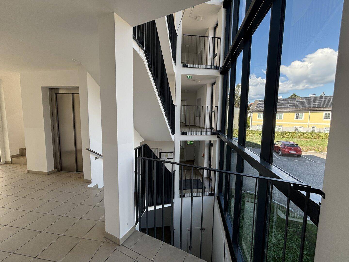 Helles Treppenhaus mit großflächiger Verglasung, Aufzug und Blick ins Freie.