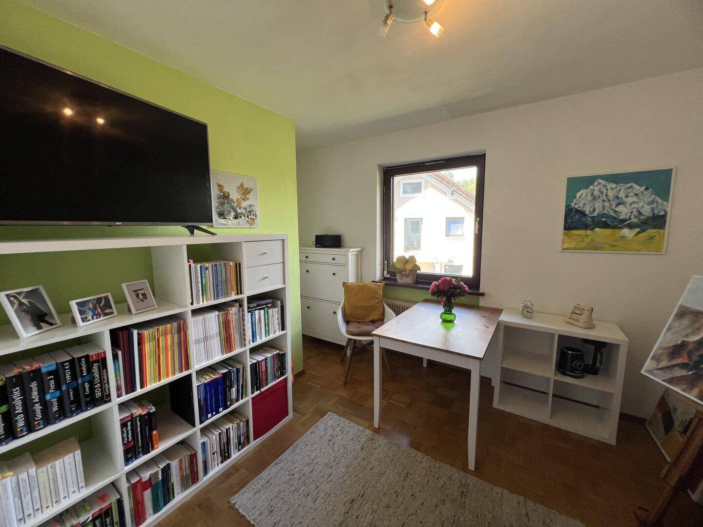 Helles Zimmer mit Bücherregal, Schreibtisch und einem Fernseher an der grünen Wand.