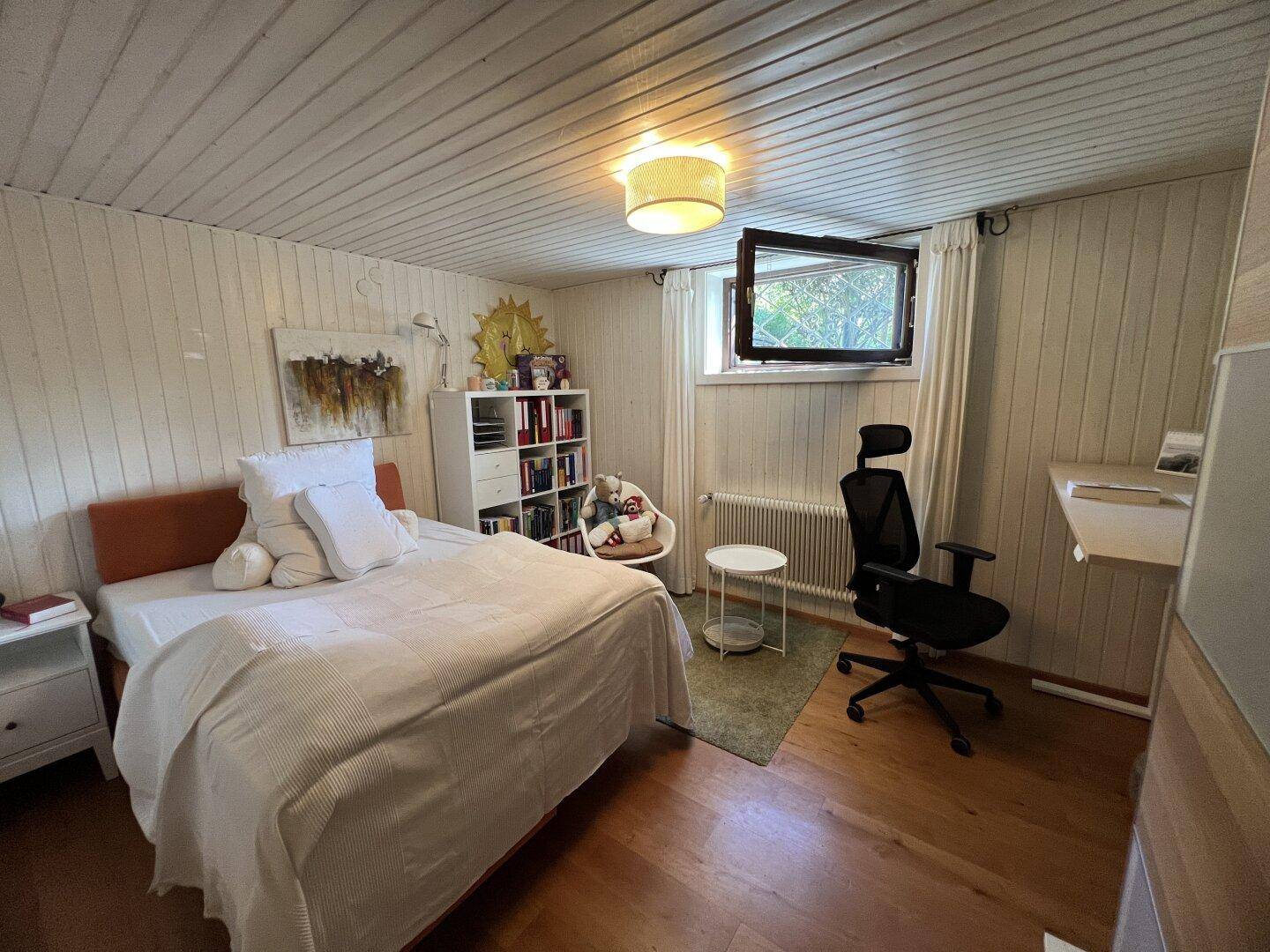 Helles Schlafzimmer mit Holzboden, weiß getäfelten Wänden, Bett und kleinem Arbeitsbereich.