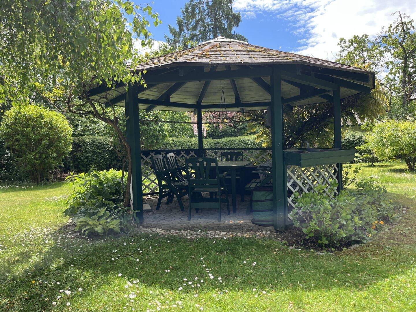 Detailansicht eines charmanten Gartenpavillons mit Tisch und Stühlen, umgeben von Grün.
