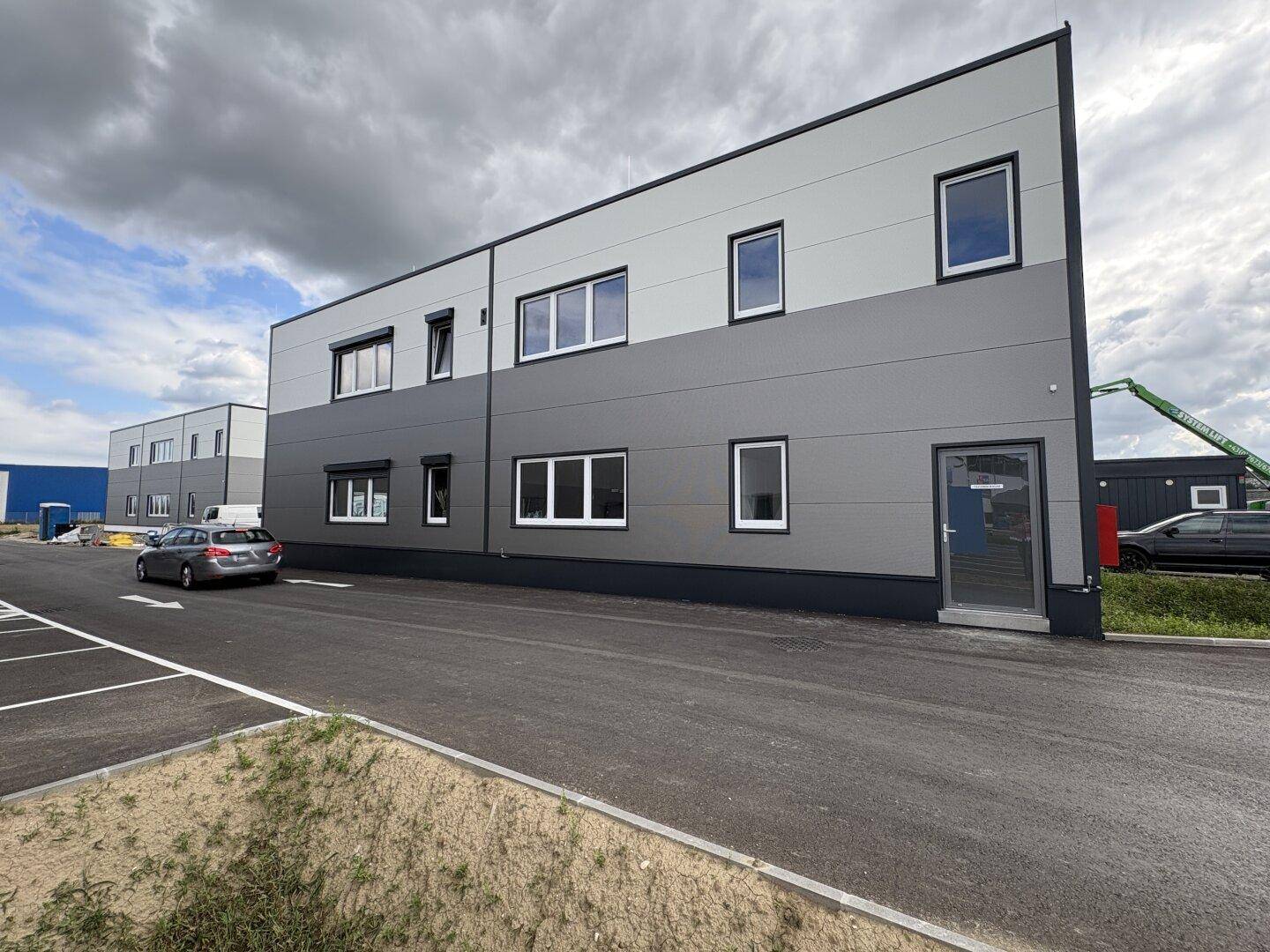 Seitenansicht eines modernen Gewerbegebäudes mit grau-weißer Fassade, vielen Fenstern und Eingangstür.