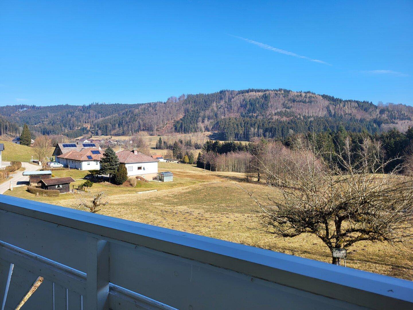 Weitblick von einem Balkon oder einer Terrasse über Felder, Wälder und entfernte Häuser unter blauem Himmel.