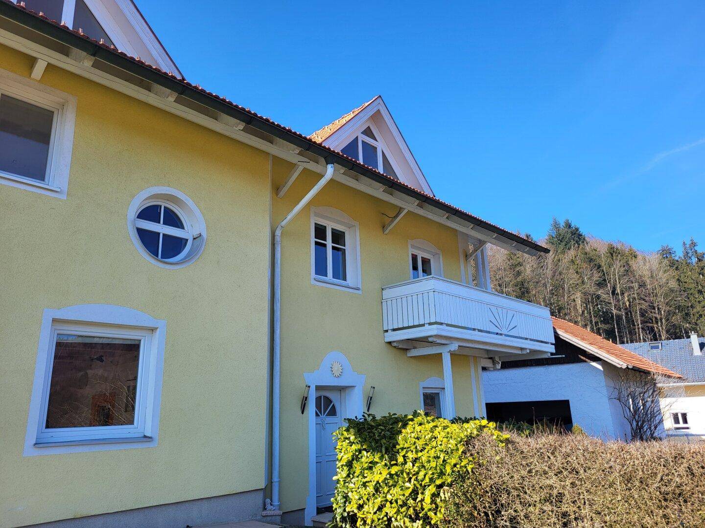 Nahaufnahme der gelben Hausfassade mit weißem Balkon und runden Fenstern unter blauem Himmel.